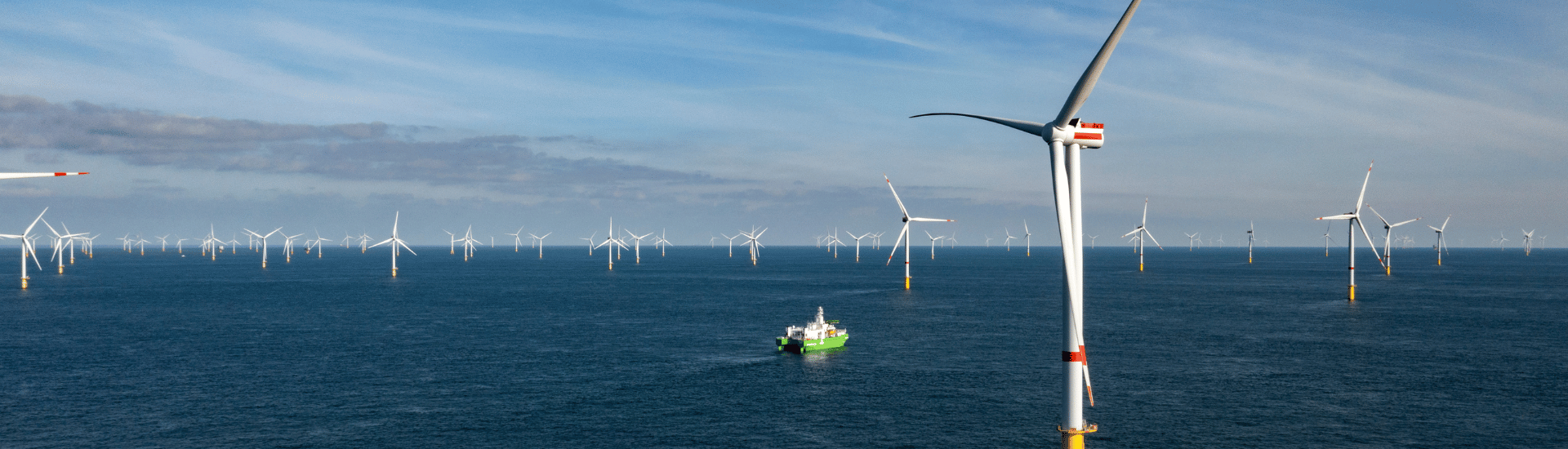 A large offshore wind farm with numerous wind turbines in the sea and a green and white ship moving through the water under a partly cloudy sky.