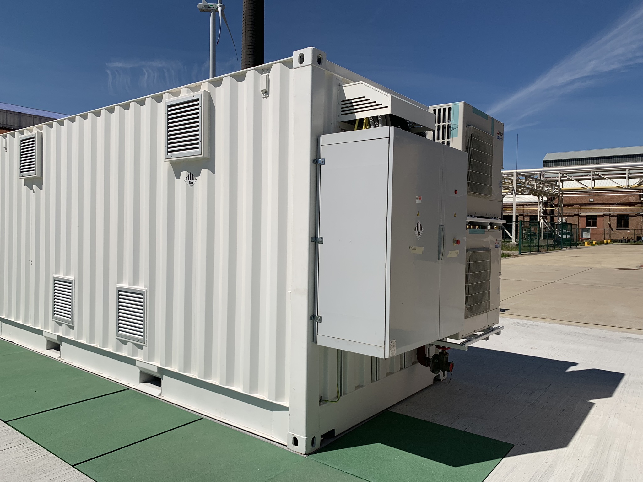 Containerized battery installation at an industrial site with a wind turbine in the background