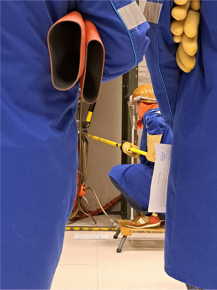 Workers in protective blue clothing handle electrical equipment with an insulated pole in an industrial setting.